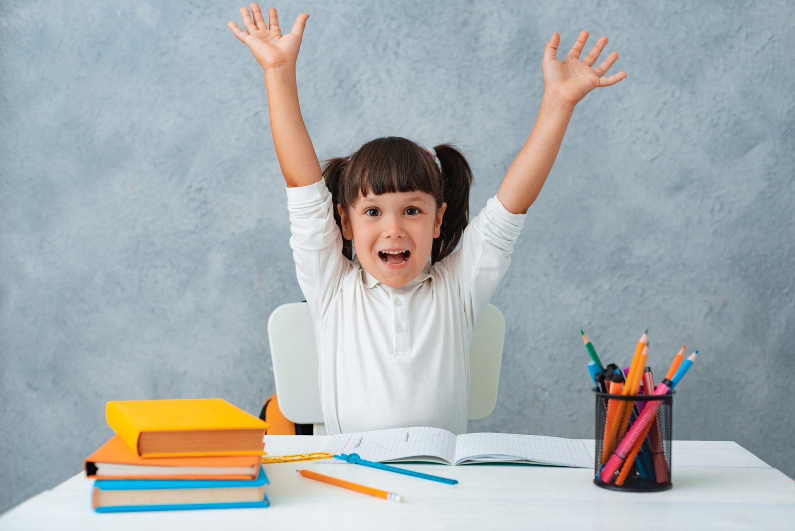 Back to school. Cute child schoolgirl sitting at a Desk in the room. The kid is learning and doing his homework. The girl is emotionally happy with her hands up